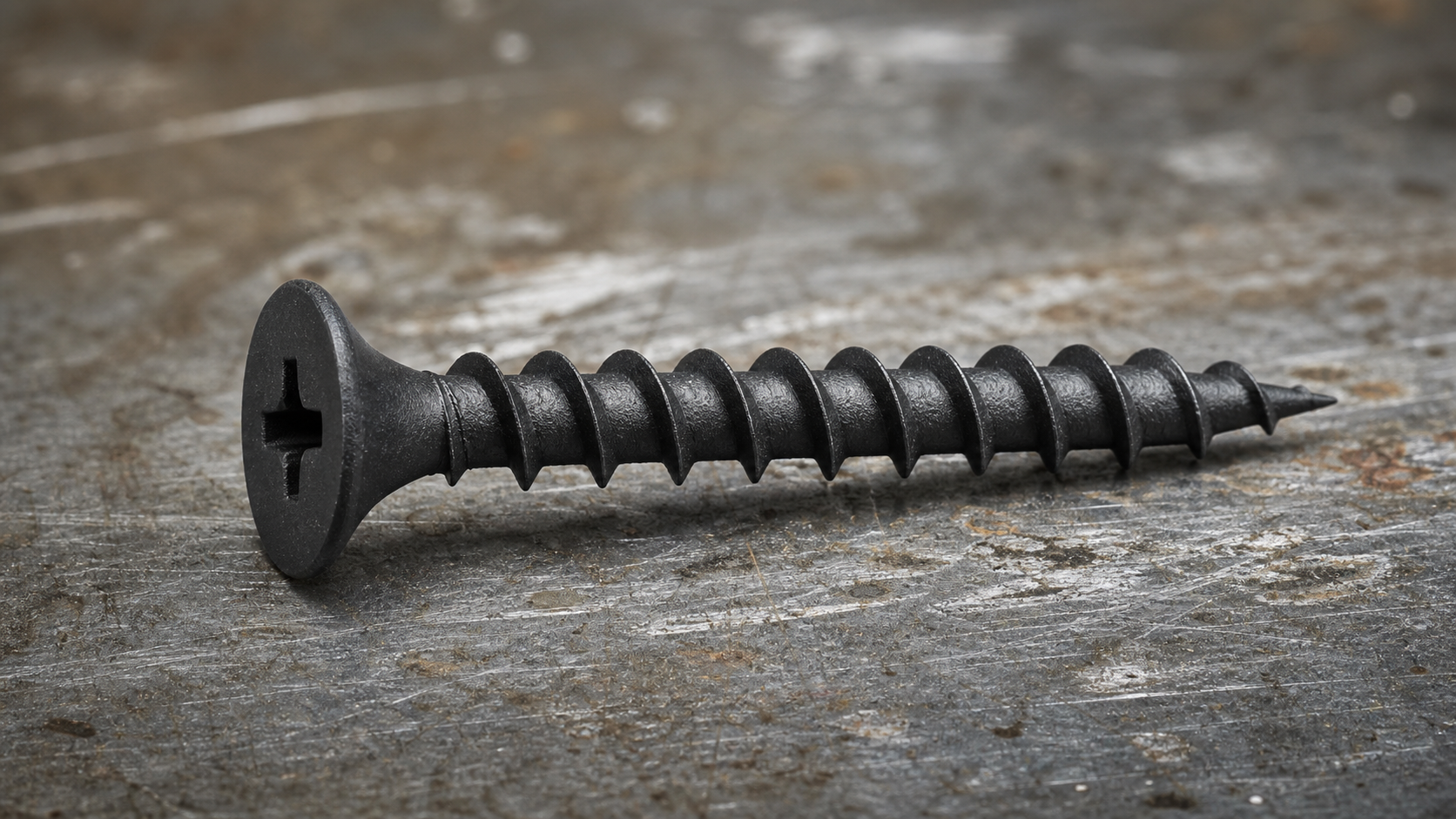 Close-up of a black phosphate drywall screw with bugle head, Phillips drive, deep coarse thread, and needle-point tip on whit